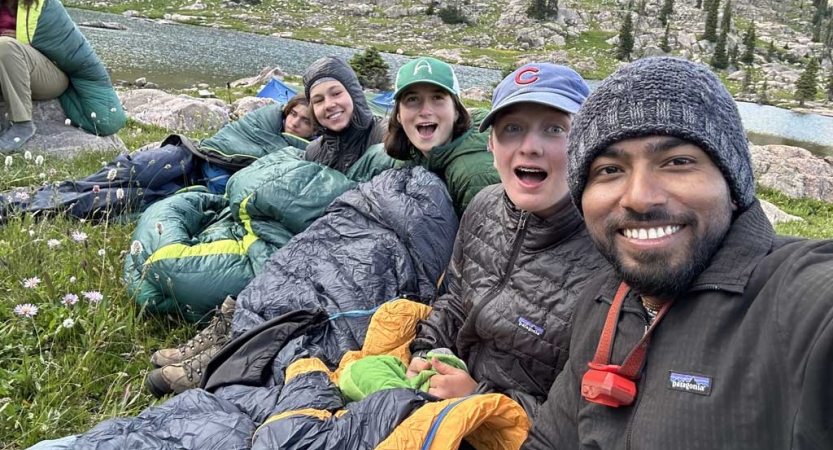 a group of people lay or sit on the ground, bundled in coats and sleeping backs and smile for the photo 